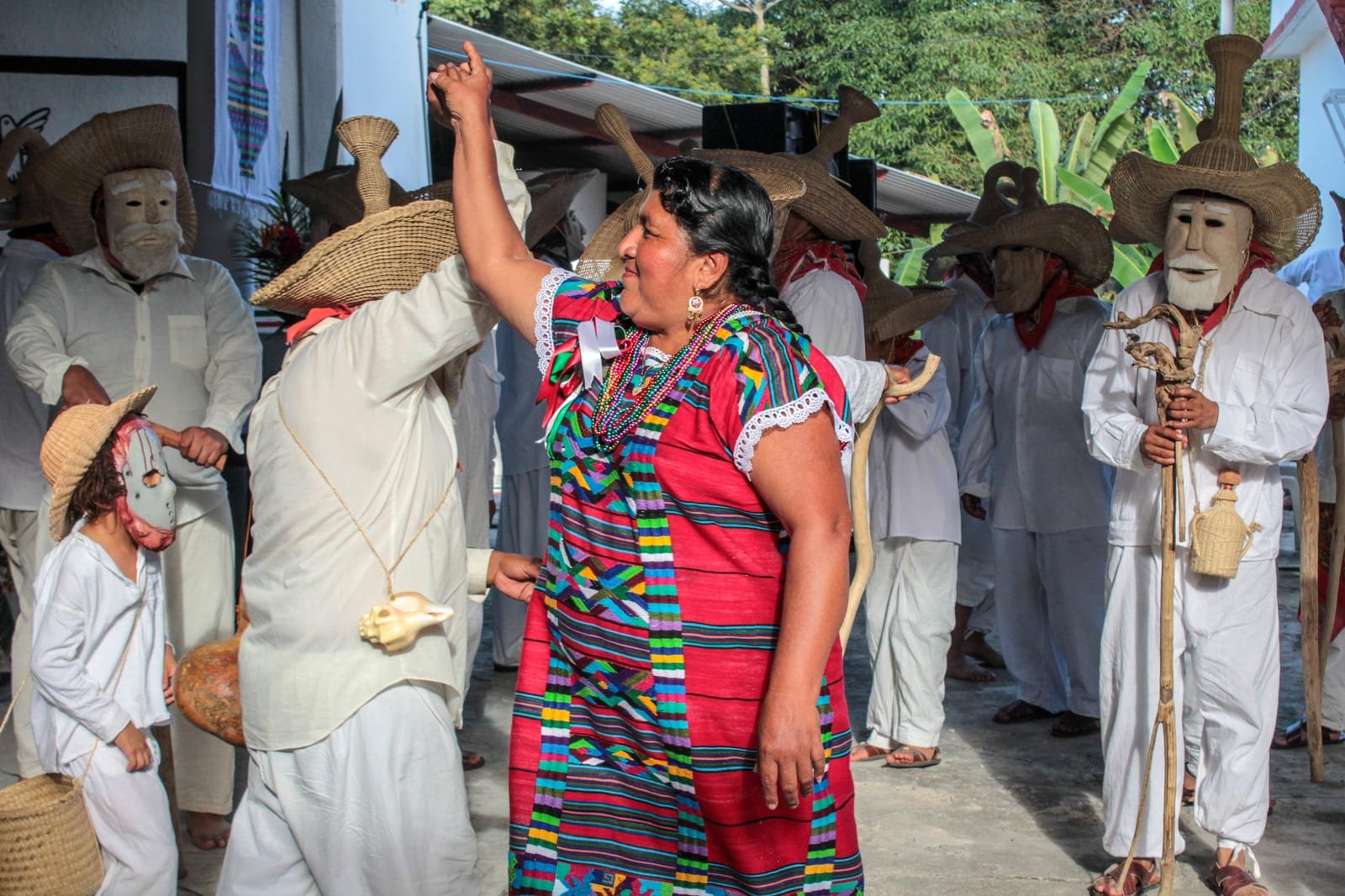 A woman in bright traditional Oaxacan dress dancing among masked huehuentón figures at a Day of the Dead festival.